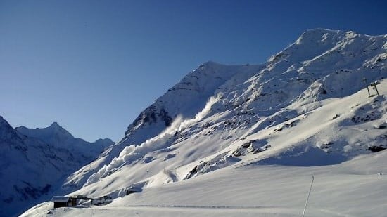 Vue des montagnes enneigées avec des avalanches en cours, sous un ciel bleu clair.
