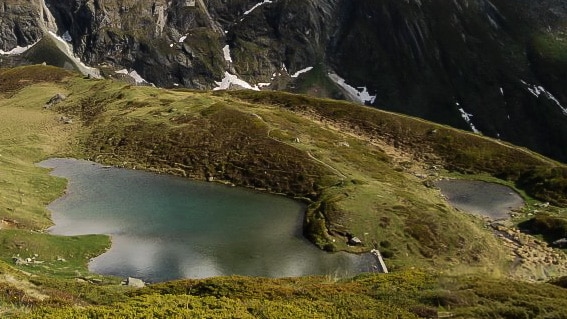 Vue panoramique du Lac d'Ar Pitetta, entouré de pelouses alpines verdoyantes et de montagnes. Le lac est peu profond et reflète le paysage environnant, avec quelques restes de neige dans les hauteurs.