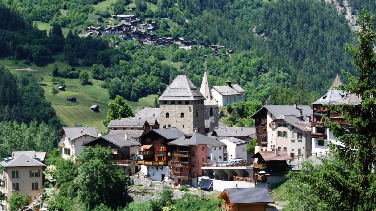 Village de montagne pittoresque avec tour et église, entouré de verdure.