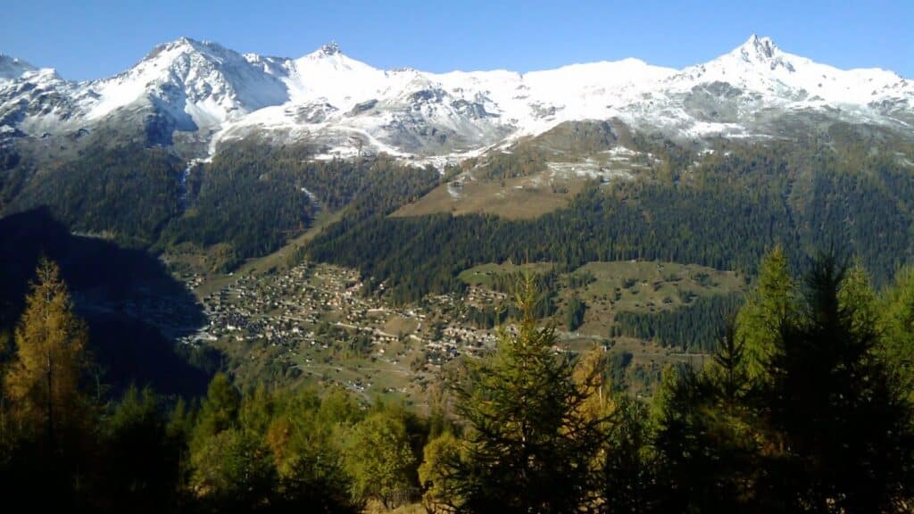 Panoramic view of a mountainous landscape in autumn, featuring snow-capped peaks and a valley with a small village surrounded by forests.