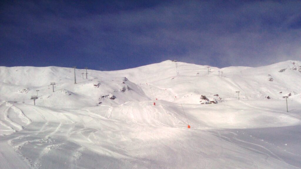 Vue d'une montagne enneigée avec des pistes de ski et des remontées mécaniques sous un ciel partiellement nuageux.
