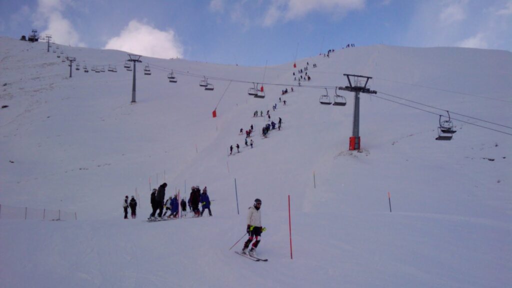 Skiers on a slalom slope with ski lifts in the background, under a partly cloudy sky.