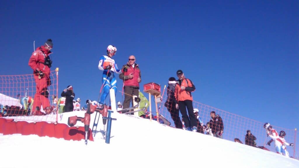 Des skieurs et des membres du personnel se préparent sur une piste de slalom géant, avec des montagnes en arrière-plan sous un ciel dégagé.