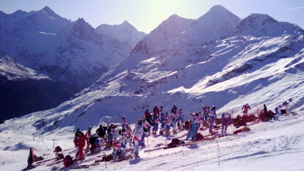 Skieurs et entraîneurs sur une piste de ski sous un ciel ensoleillé, avec des montagnes enneigées en arrière-plan.