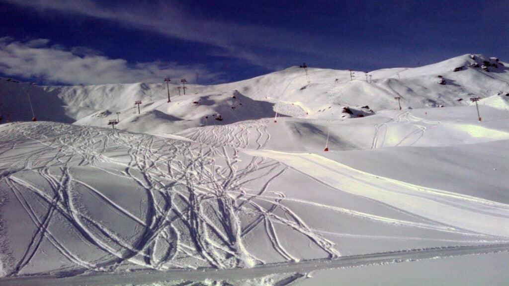 Vue panoramique d'une station de ski avec des pentes enneigées, des traces de ski sur la neige, et des remontées mécaniques dispersées sur fond de ciel partiellement nuageux.