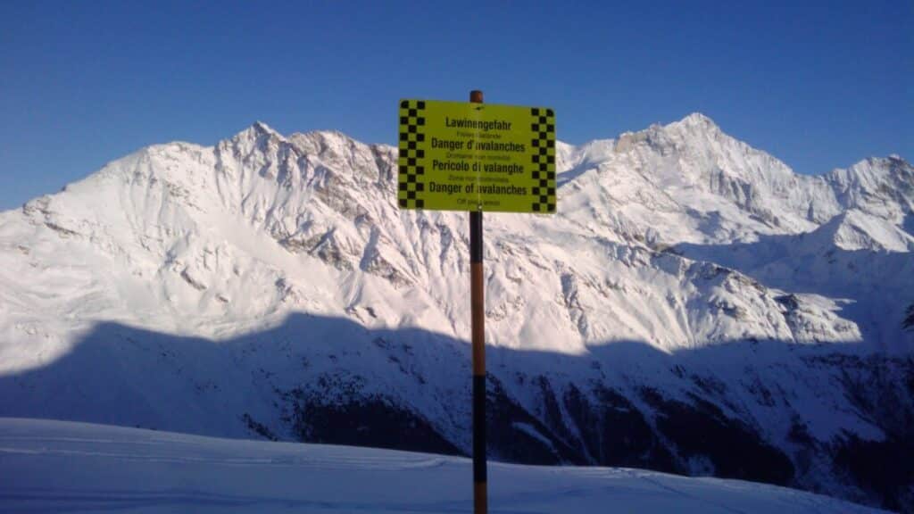 A warning sign indicating avalanche danger in a snow-covered mountain landscape under a clear blue sky.