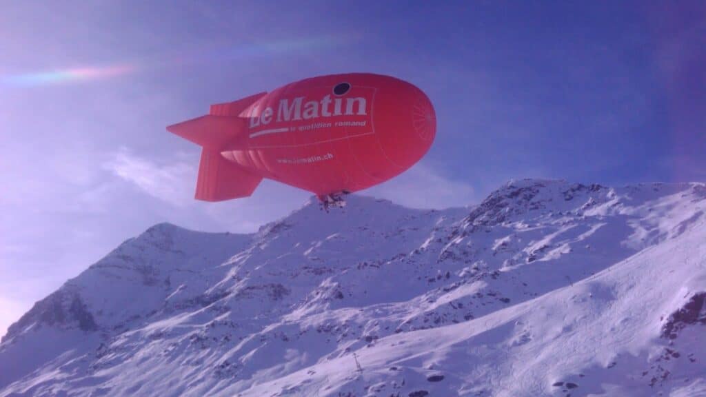 Un dirigeable rouge de "Le Matin" survole des montagnes enneigées sous un ciel bleu clair.