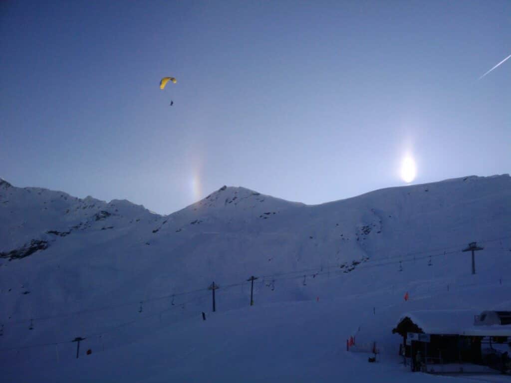 Paraglider soaring over snow-covered mountains with a bright sun and rainbow-like effects in the sky.