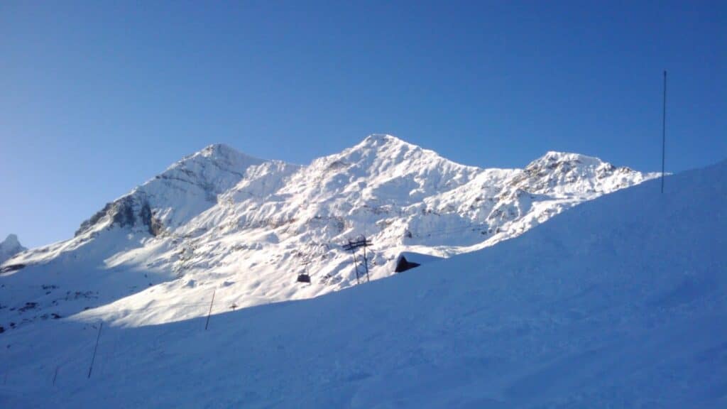 Vue des montagnes enneigées au lever du soleil, avec un ciel bleu clair et des pistes de ski en contrebas.