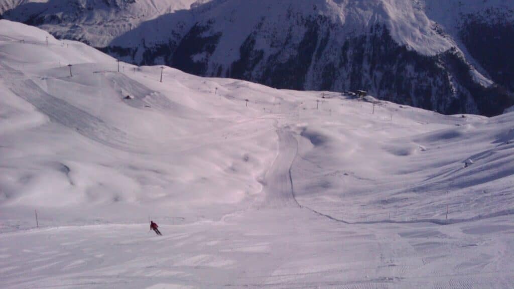 Panoramic view of snow-covered ski slopes with a lone skier at the forefront, surrounded by mountains under clear skies.