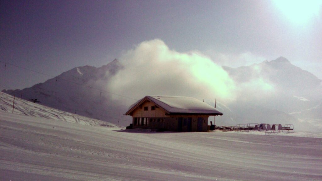 Un chalet en montagne recouvert de neige, avec des sommets montagneux en arrière-plan et des nuages s'élevant au-dessus.