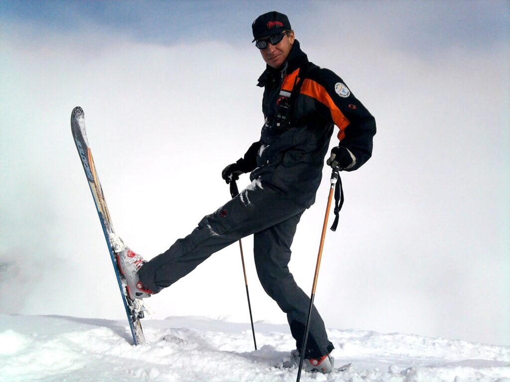 Un skieur en tenue de ski, avec des skis et des bâtons, pose dans la neige sur un fond de ciel nuageux.