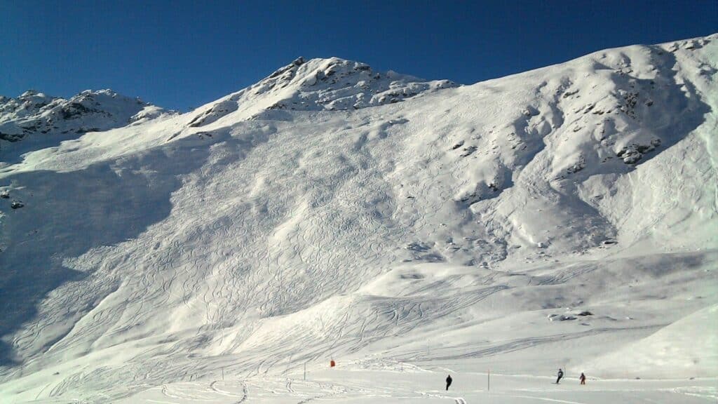 Vue panoramique d'une montagne enneigée avec des traces de skis visibles sur la pente, sous un ciel ensoleillé.