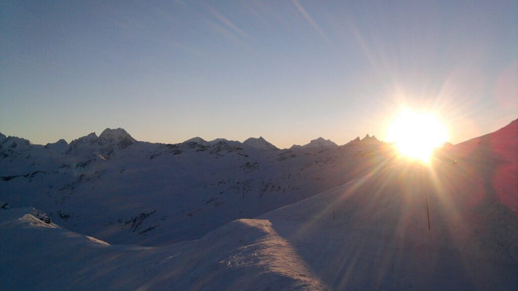 Sunset over snow-covered mountains, with the sun shining brightly on the horizon.