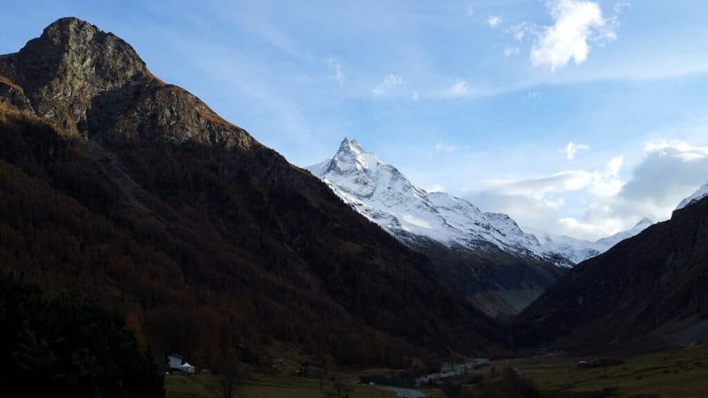 Vue panoramique des montagnes avec des sommets enneigés sous un ciel partiellement nuageux, représentant le paysage des Alpes. La vallée est entourée de pentes boisées et de formations rocheuses.