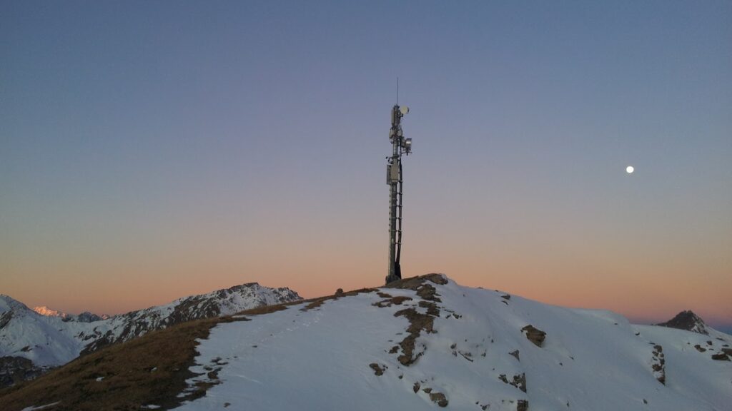 A telecommunications tower stands on a snow-covered mountain ridge during sunset, with a clear sky and the moon visible in the background.