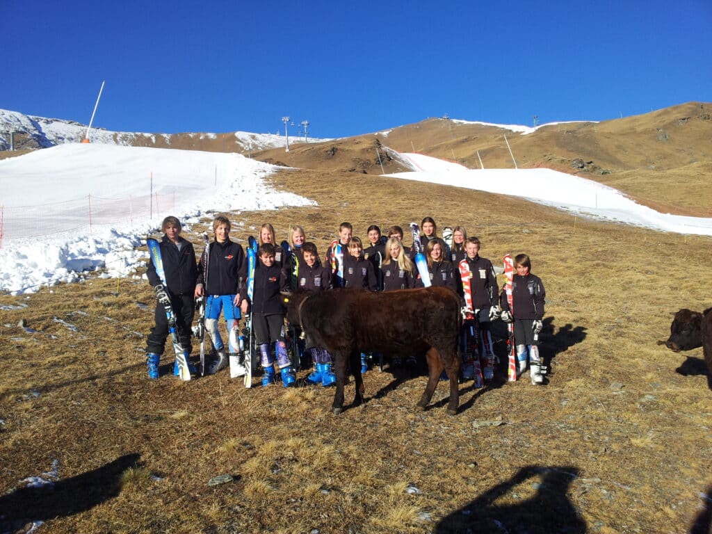 Un groupe d'enfants en ski pose au milieu d'un terrain de montagne, avec des pistes de ski en arrière-plan et des vaches Hérens à proximité.