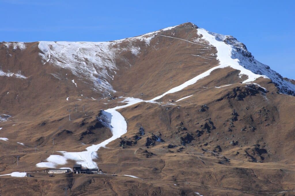 A mountainous landscape with a snow-capped peak and patches of snow on the slopes, featuring a ski area with a lodge and ski lifts.