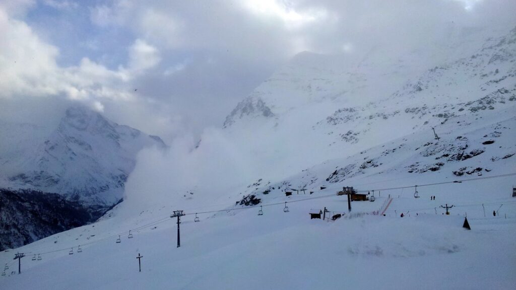 Vue d'une station de ski avec des montagnes enneigées en arrière-plan et des remontées mécaniques visibles sous un ciel nuageux.