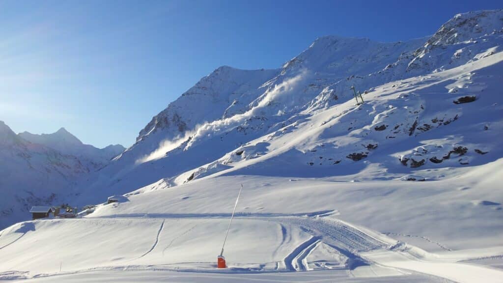 Vue panoramique d'un paysage de montagne enneigé avec des pistes de ski, un bâtiment au premier plan et des sommets majestueux sous un ciel bleu clair.
