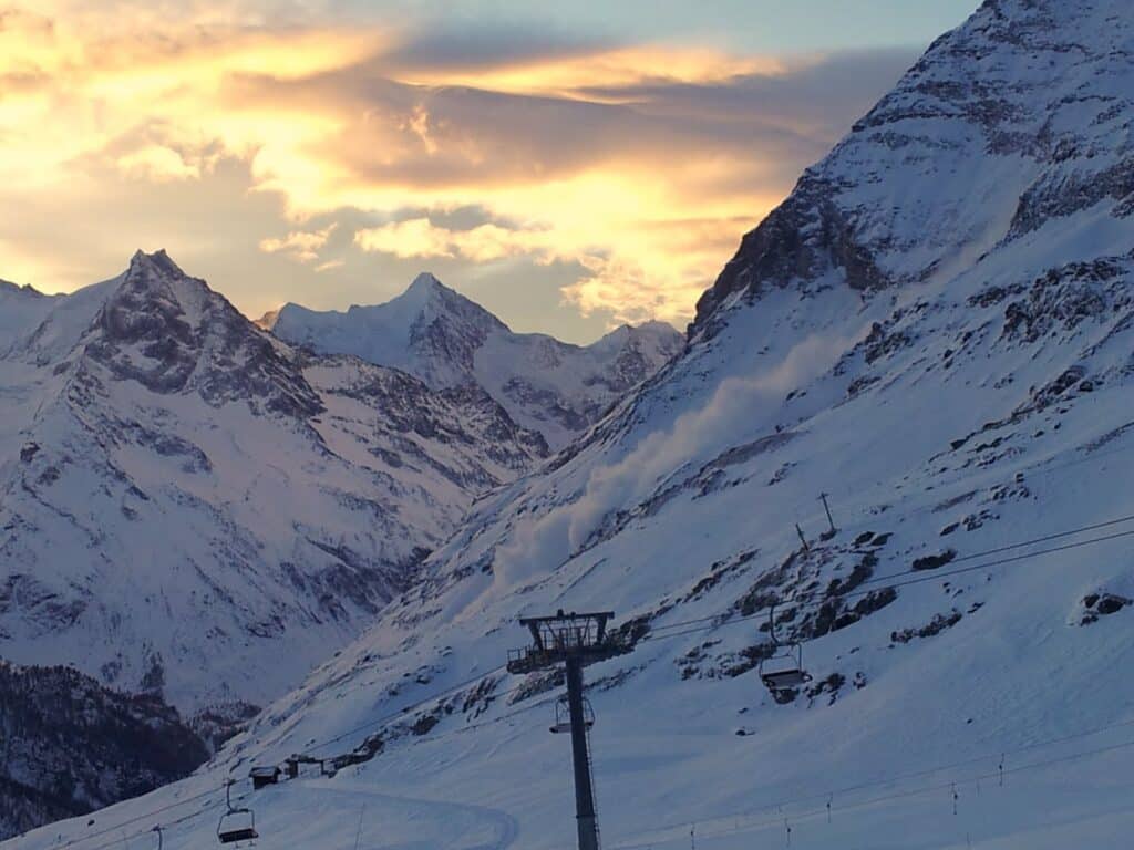 Vue des montagnes enneigées avec un nuage de neige soulevé et un télésiège en bas, sous un ciel coloré au coucher du soleil.