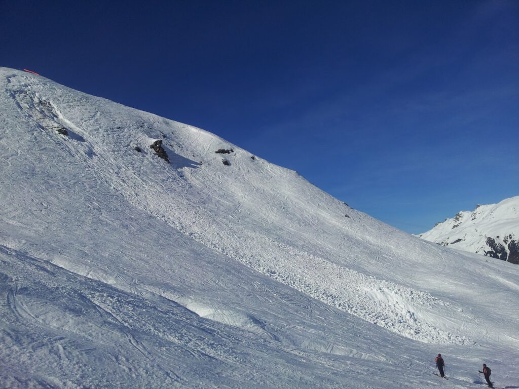 Vue d'une pente enneigée avec des traces de ski, sous un ciel bleu clair, deux skieurs marchent sur la neige.