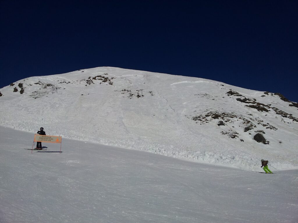 Un skieur dans une pente enneigée, avec des panneaux de sécurité en avant-plan et un sommet de montagne couvert de neige en arrière-plan.