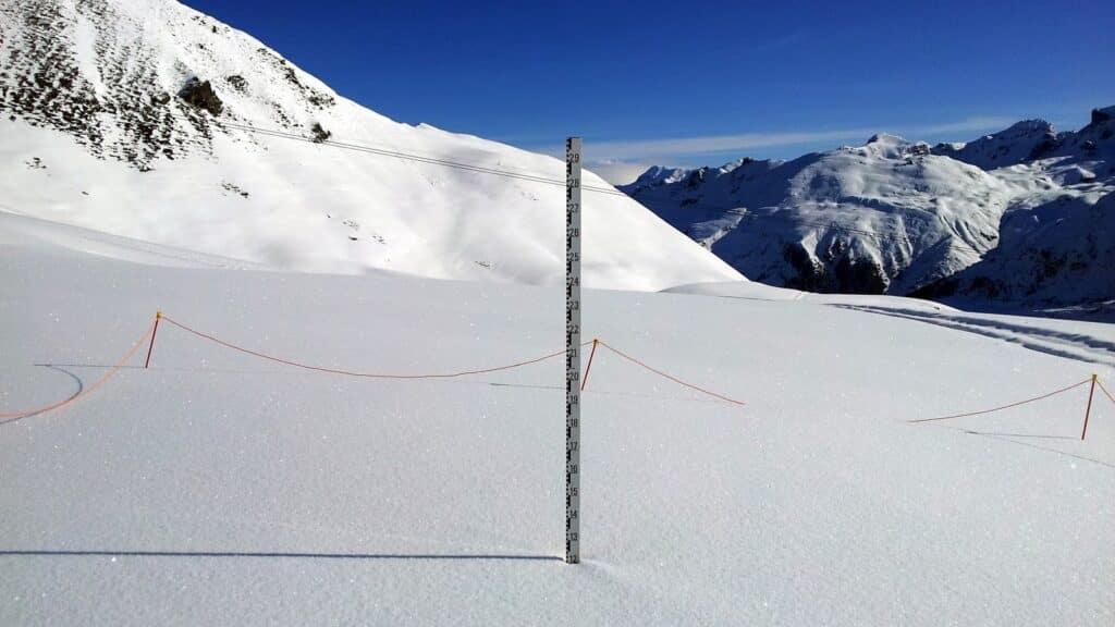 A snowy mountain landscape with a measurement pole indicating snow depth, surrounded by skiing barriers and distant peaks under a clear blue sky.