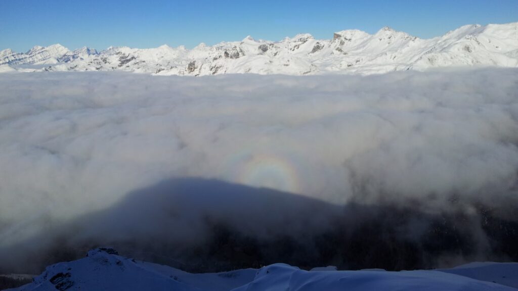 Un halo lumineux émerge au-dessus d'une mer de brouillard, avec des montagnes enneigées en arrière-plan.