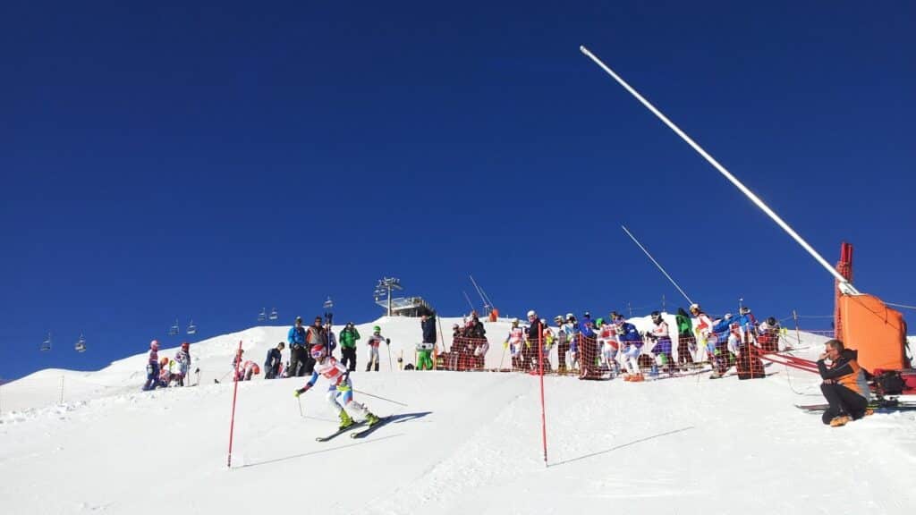 Une athlète en train de skier sur une piste de slalom, entourée de spectateurs et de compétiteurs au départ, sous un ciel bleu.