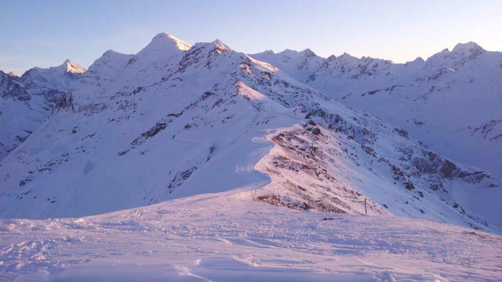 Vue panoramique des montagnes enneigées avec des crêtes et des pentes escarpées, capturée au coucher du soleil. Le paysage est illuminé par des teintes dorées et violettes.