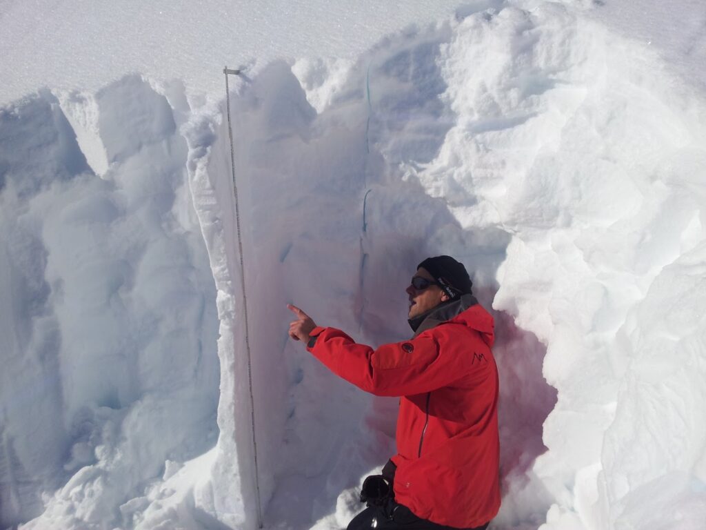 Un homme en manteau rouge examine et indique quelque chose à l'intérieur d'un profil de neige, dans un environnement enneigé.