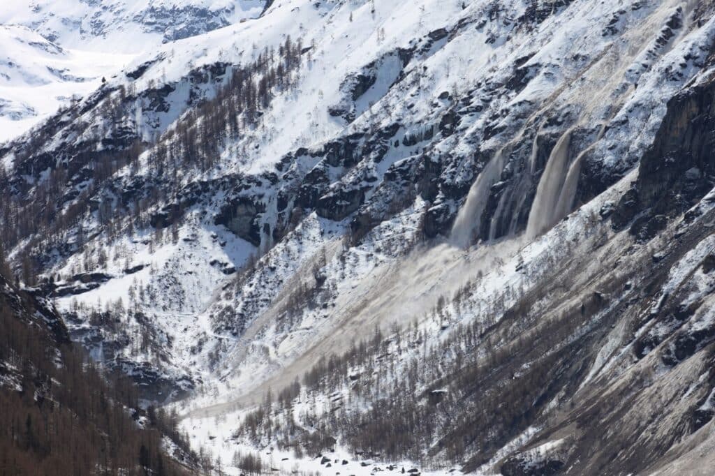 Vue d'une avalanche récente avec de la neige et des débris glissant sur une pente montagneuse, entourée de forêts et de pics enneigés.