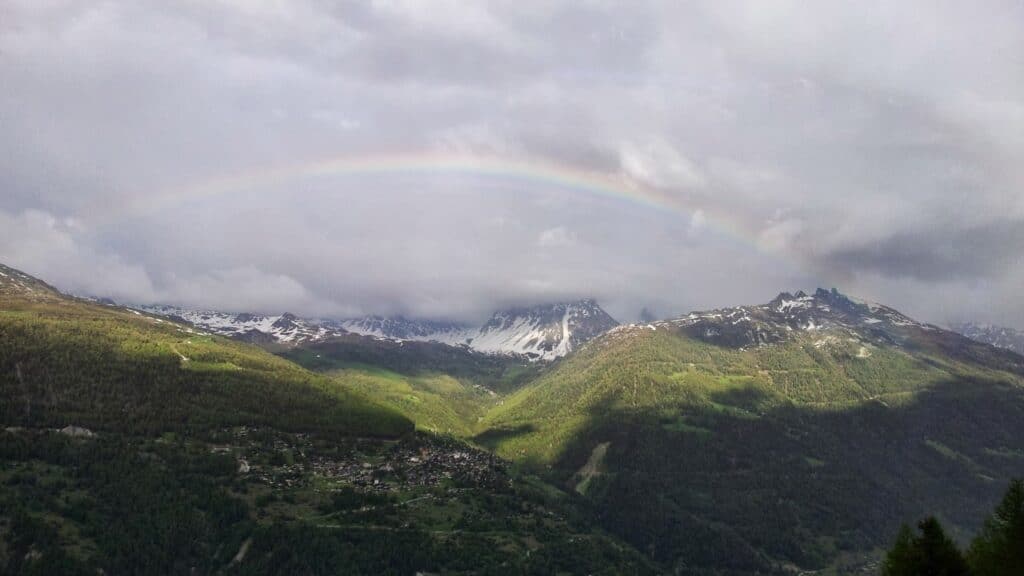 Un arc-en-ciel vibrant s'étend au-dessus des montagnes enneigées, entouré de nuages sombres et de verdure, capturant un moment magique après la pluie.