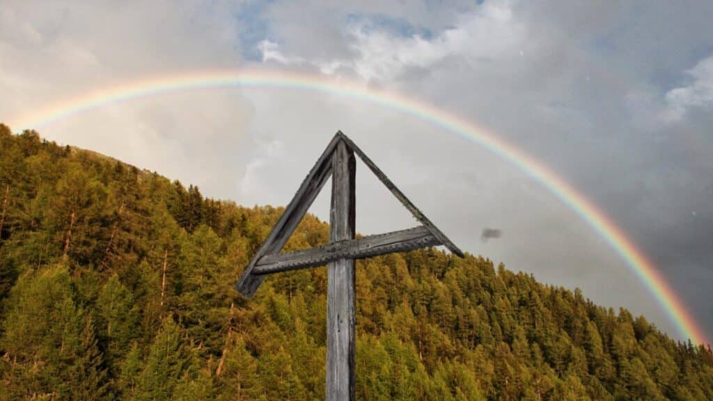 Un arc-en-ciel s'étend au-dessus d'une forêt de sapins, avec une croix en bois au premier plan.