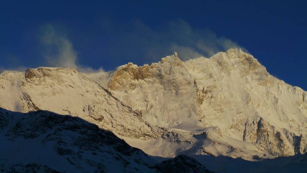 Vue du sommet enneigé d'une montagne sous un ciel bleu, avec des nuages de vent soufflant au sommet.