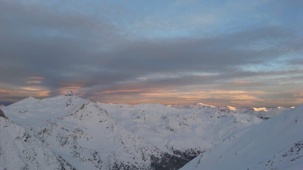 Vue panoramique des montagnes enneigées au lever du jour, avec un ciel nuageux et des teintes pastel à l'horizon.