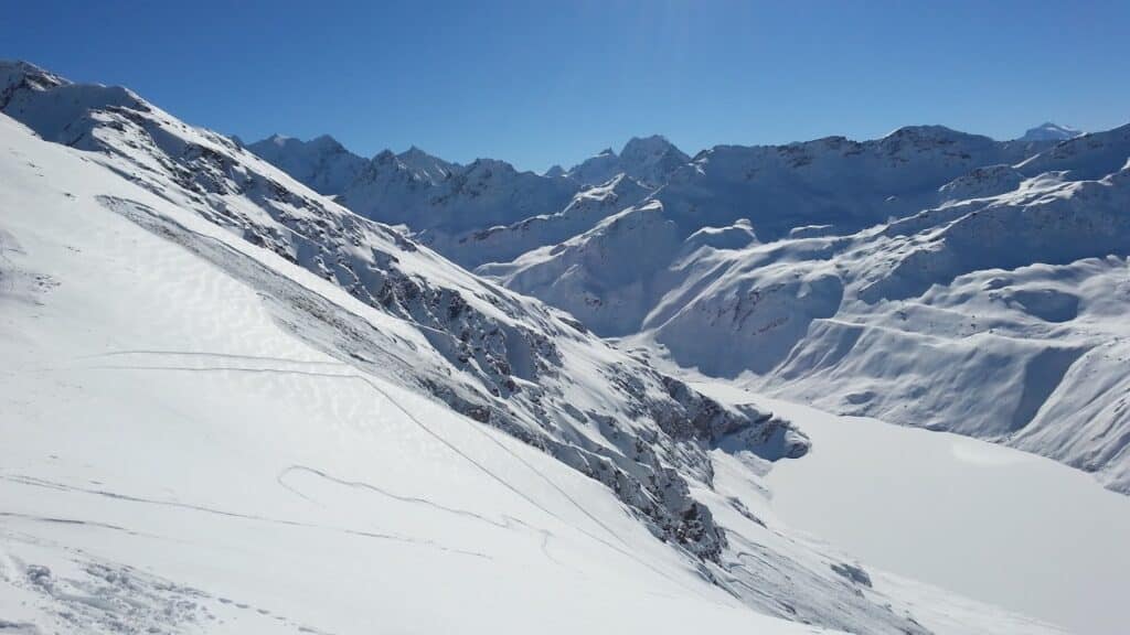 Vue panoramique des montagnes enneigées avec un lac en bas, capturée sous un ciel ensoleillé.