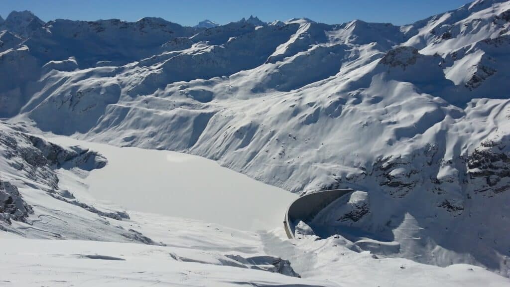 Vue panoramique des montagnes enneigées avec un lac gelé au premier plan, sous un ciel bleu dégagé.