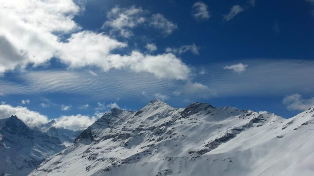 Vue panoramique des montagnes enneigées sous un ciel partiellement nuageux, avec des nuages blancs et des crêtes montagneuses recouvertes de neige brillante.