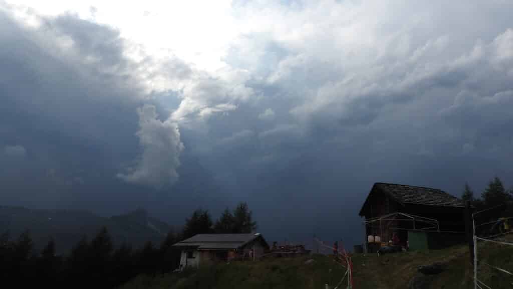 Vue d'un paysage montagneux avec des nuages sombres, un chalet en bois au premier plan, et des arbres sur le côté.
