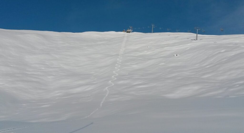 Vue d'un large terrain de neige avec une trace de ski marquée sur la surface, sous un ciel bleu dégagé.