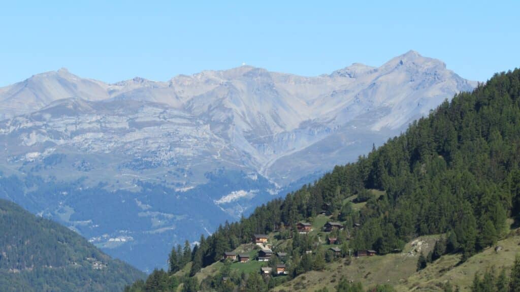 Vue panoramique des montagnes avec des chalets en bois au premier plan et des pentes verdoyantes.