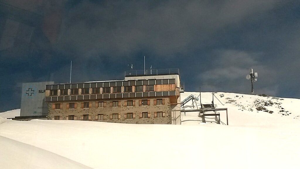 Vue du bâtiment du SLF (Institut Fédéral pour l'Etude de la Neige et des Avalanches) recouvert de neige, avec un ciel partiellement nuageux en arrière-plan.