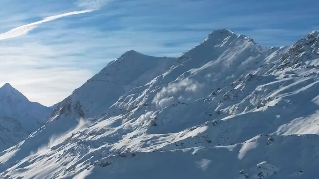 Vue des montagnes enneigées avec des nuages légers et des traces de neige en mouvement sur les pentes, capturée par une belle lumière matinale.