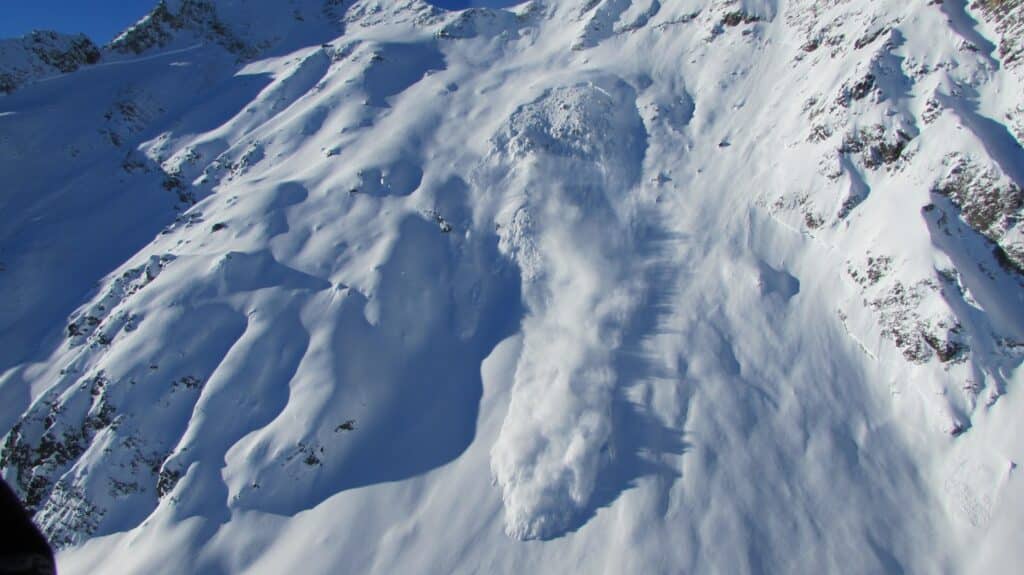 Vue aérienne d'une avalanche de neige descendante sur une pente enneigée, montrant des couches de neige et des rochers environnants.