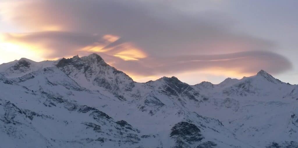 Vue des montagnes enneigées sous un ciel coloré au crépuscule, avec des nuages lenticulaires au-dessus des sommets.