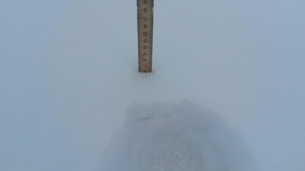 Mètre de mesure enfoncé dans la neige, affichant une lecture de 10 cm sur un fond de neige blanche.