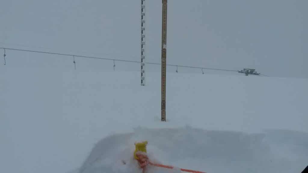 Panneau de mesure de la neige dans un environnement enneigé, indiquant une forte accumulation de neige, avec des pylônes de remontée en arrière-plan.