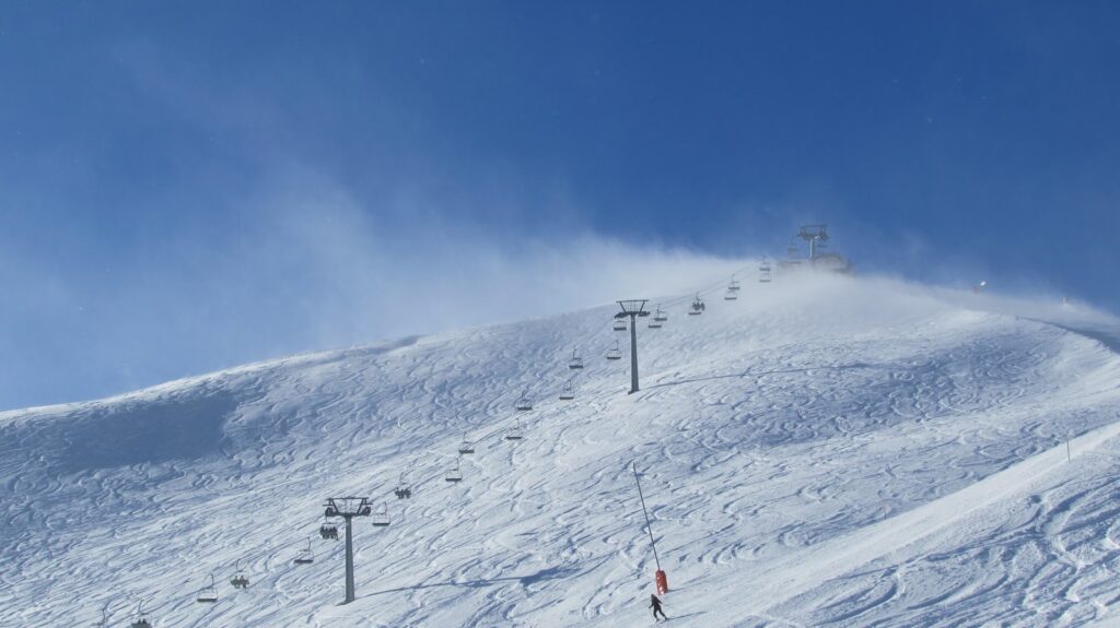 Un skieur sur une pente enneigée avec des télésièges visibles sous un ciel bleu clair et venteux.